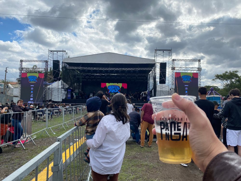 Sob o céu azul oculto por pesadas nuvens carregadas, um palco de festival se ergue em uma praça lotada de pessoas. O letreiro colorido em vermelho, amarelo e azul traz as palavras “Orbital Rock”. De frente à cena, uma mão se ergue segurando um copo pela metade de cerveja dourada com a estampa “Cervejaria Pangea”.