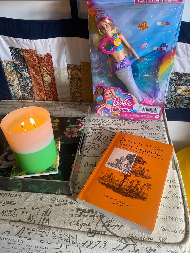 An orange book in front of a multi colored blond haired mermaid Barbie on a tan and brown table with a green and white candle