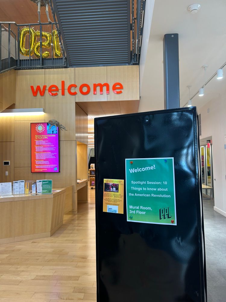 Entrance to a building with light wood floors, a black poster display and a bright orange banner that says welcome 