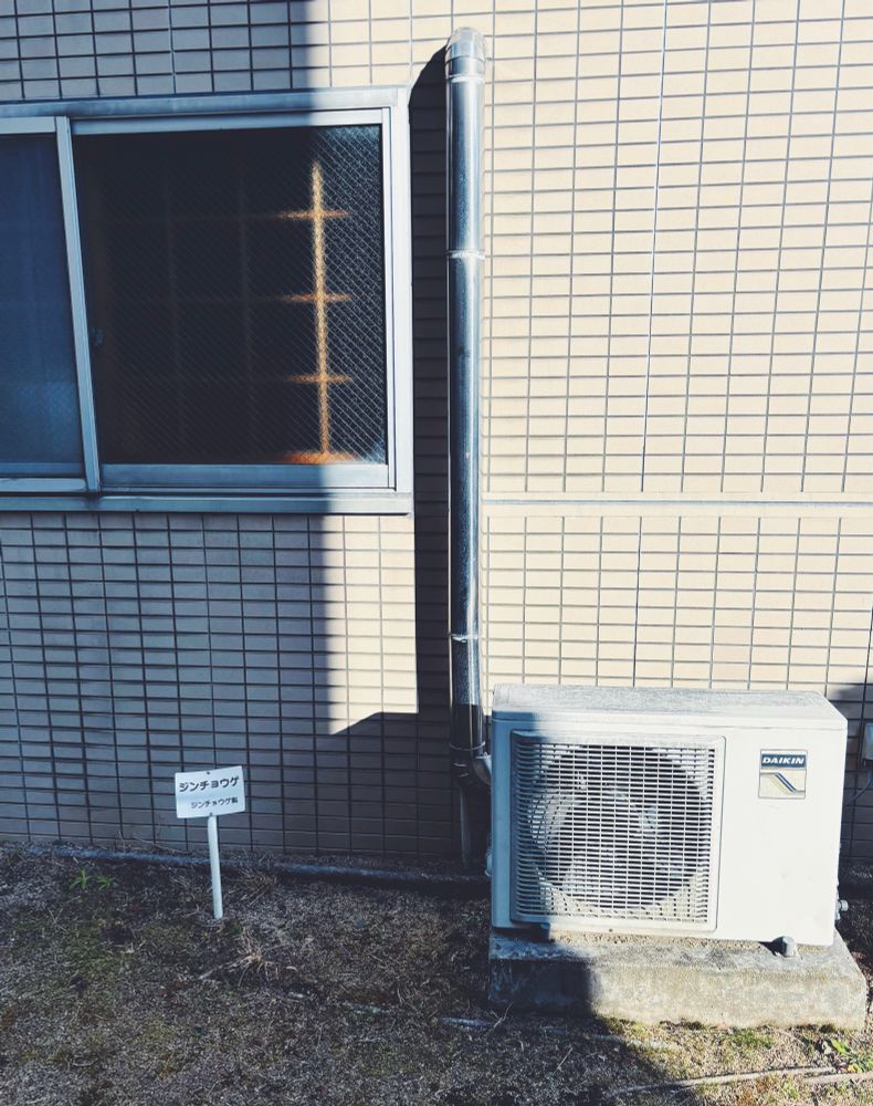 ダイキン製のエアコンの室外機の側に、「ジンチョウゲ ジンチョウゲ科」という札が立てられている  ジンチョウゲはどこにもない