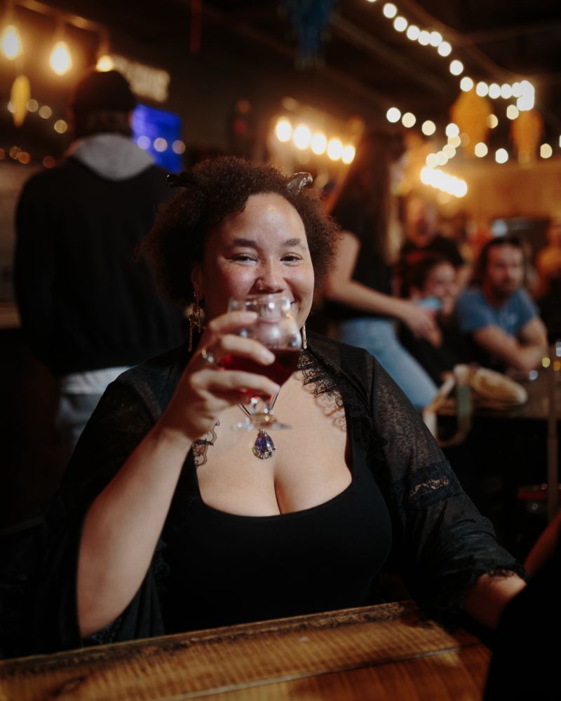 A woman wearing black clothing and satyr horns drinks wine in a brewery lit by string lights. 