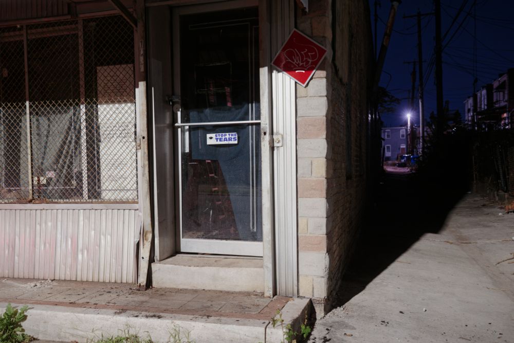 An abandoned formstone storefront with a bumper sticker on the glass door that says "STOP THE TEARS."
