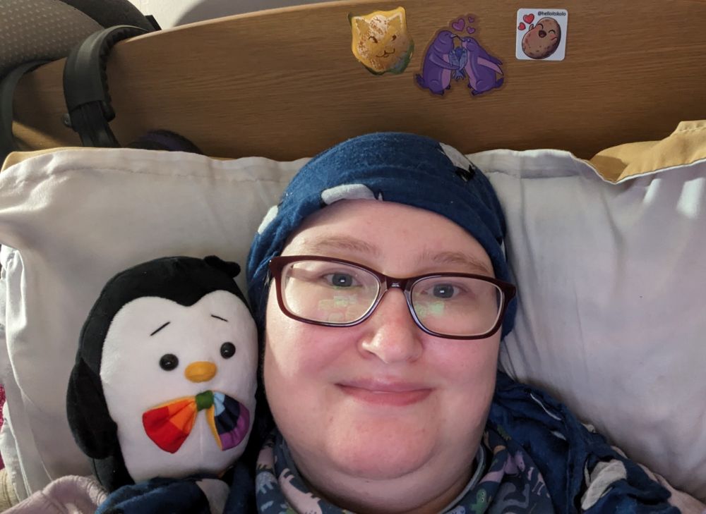 Danni, a white person, lying in bed, smiling. They're wearing purple glasses, a blue penguin headscarf, and a dinosaur bib. On their shoulder is a soft toy penguin, which has a rainbow bow tie. 