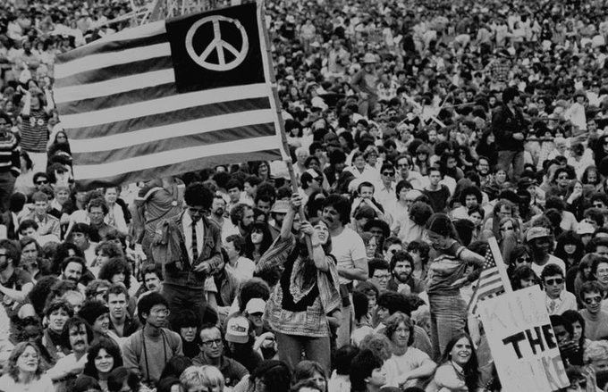 A black and white photograph of the some of the tightly-packed crowd at the rally on the Great Lawn in Central Park. Near the center, a woman stands up holding an American flag whose stars have been replaced with the peace symbol.