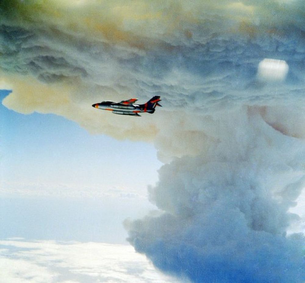 A close-up color aerial photograph of the large white mushroom cloud from the Canopus test with a fighter jet (probably conducting a dangerous air sampling mission to collect radioactive debris in order to analyze the efficiency of the explosion) in the foreground.