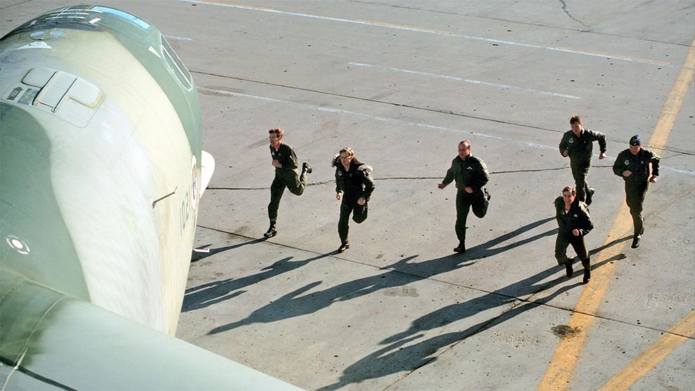A color photograph of the six-man crew of a B-52 bomber runs across the apron toward their aircraft during an alert exercise. The photograph appears to be taken from atop the right wing of the plane, which is painted in camouflage colors.