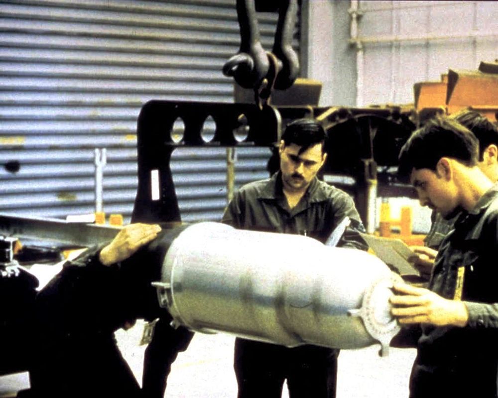 A color photograph of a team of four US Air Force airmen handling a silver-colored W80-1 thermonuclear warhead inside a maintenance facility. One man has his left hand on the front of the warhead while the man next is reading something, probably a checklist.