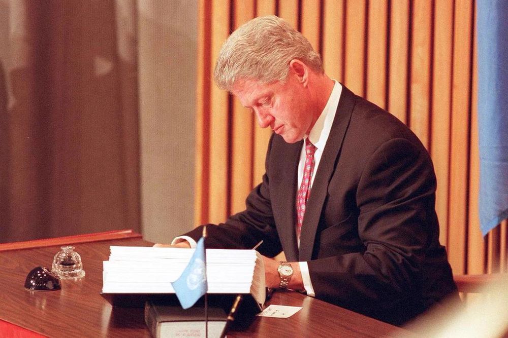 A color photograph of President Bill Clinton signing the Comprehensive Nucear-Test-Ban Treaty at a desk during a ceremony at the United Nations on September 24, 1996. A small UN flag is on the desk in the foreground.