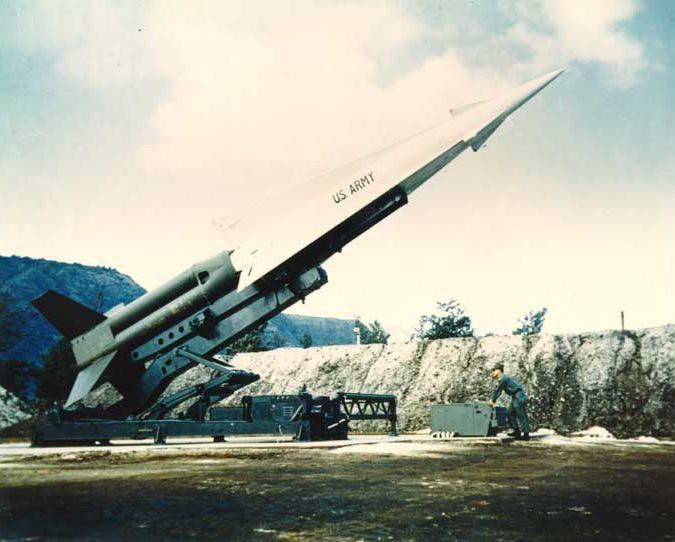 A color photograph of a Nike Hercules anti-aircraft surface-to-air missile at an Army base. The white missile is mounted on a green rail launcher and titled at about a 45-degree angle toward the partly cloudy sky. A soldier in a green uniform at right leans on a large green metal box adjacent to the launcher.