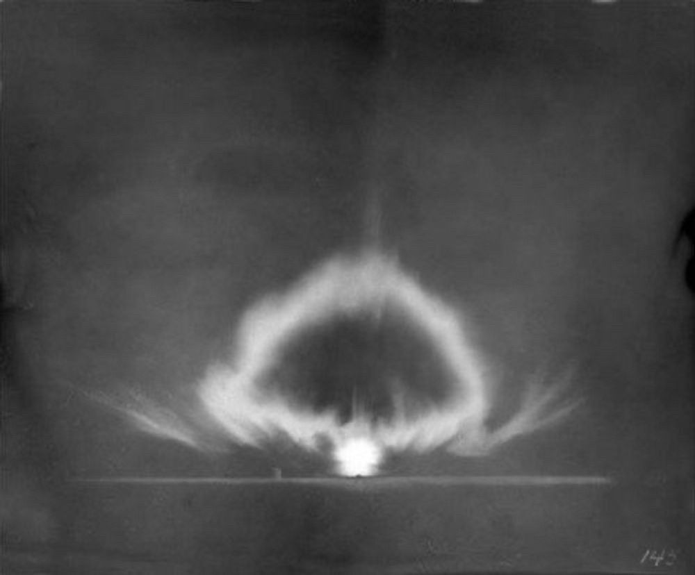 Another black and white photograph of the Trinity explosion a few milliseconds later, showing an almost ghostly mushroom cloud with glowing tendrils rising from the ground above the very bright fireball.