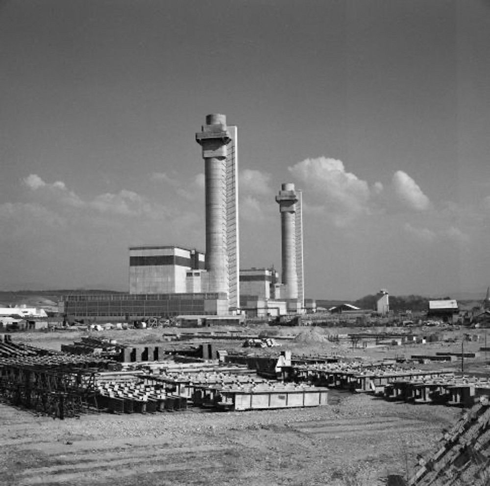 A black and white photograph of the Windscale Piles, including the two prominent and tall chimneys (one for each reactor). The large, square protrusions near the top were known, until the fire, as Cockcroft’s Folly after physicist John Cockcroft who, as director of the Atomic Energy Research Establishment, insisted (following the recommendation of fellow physicist Terence Price) that high-performance air filters be installed in the chimneys to prevent deadly radioactive particles from escaping into the atmosphere in case an aluminum-clad uranium fuel cartridge should rupture during routine operations and catch fire.