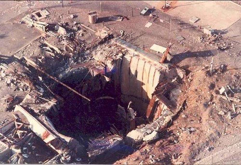 A low-level aerial color photograph of the remains of launch complex 374-7 in the aftermath of the explosion. The massive silo door is gone and debris is strewn around the area.