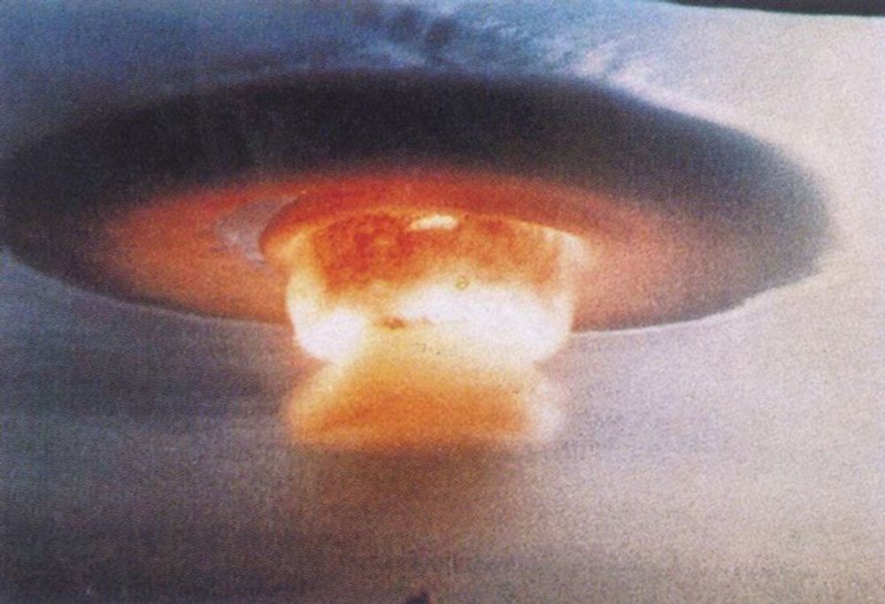 A color photograph showing the slightly later formation of the large orange-yellow mushroom cloud created by the explosion of China’s first thermonuclear weapon, with a circular condensation cloud surrounding the upper layer of the cloud.