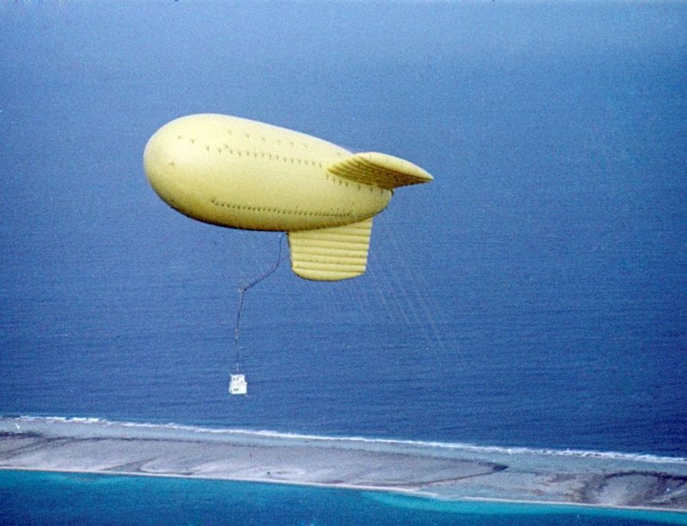 A color aerial photograph of yellow tethered dirigible-shaped balloon above Fangataufa Atoll. The relatively small white shot cab holding the thermonuclear device can be seen hanging below the balloon. Multiple thin, white tethers from the balloon to the ground are also visible.