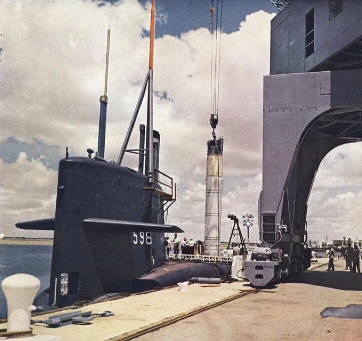 A color photograph of the USS George Washington dockside as a Polaris SLBM is lowered from a large crane into a launch tube on its hull. "598" is painted in white on its sail. Men standing on the submarine and the dock provide a sense of scale.