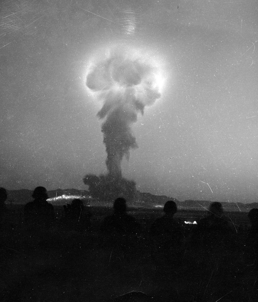 A black and white photograph of the same scene a few second later, now zoomed in on the mushroom cloud (whose top is still glowing from within) ascending into the sky, with the silhouetted helmeted heads of seven of the soldiers visible in the foreground.