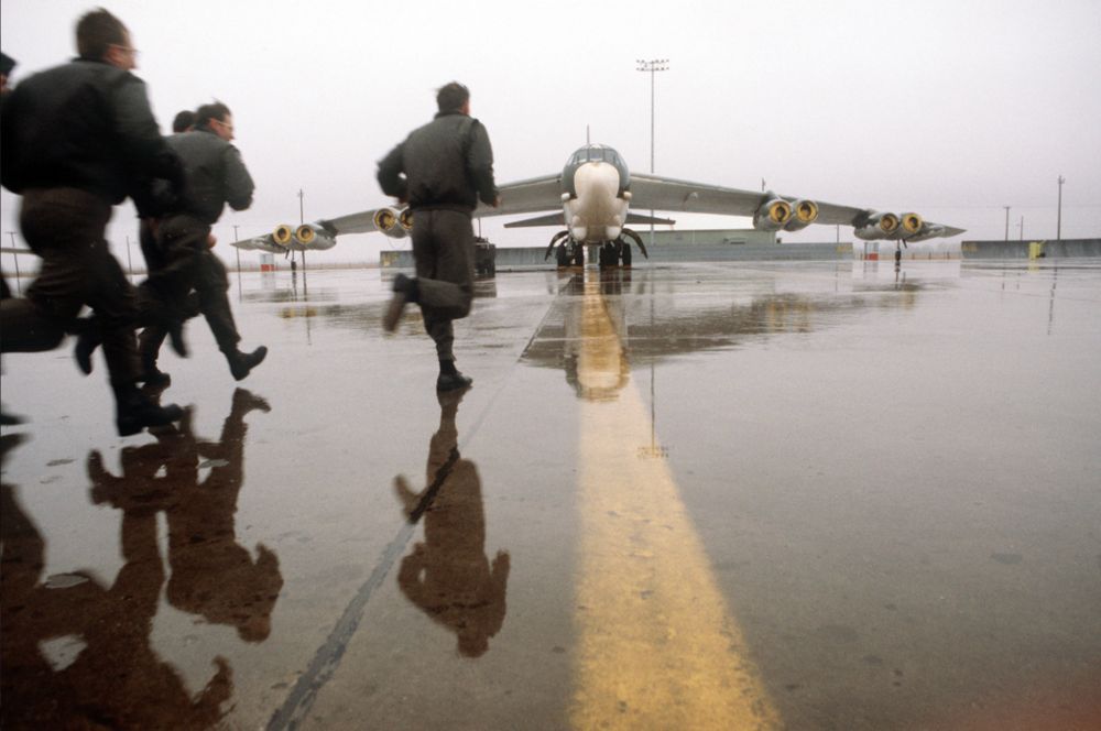 A color photograph of the crew of a B-52 strategic bomber runs across a wet apron toward their aircraft during an alert exercise at Grand Forks Air Force base, North Dakota, with both their figures and the plane reflected on the wet pavement.