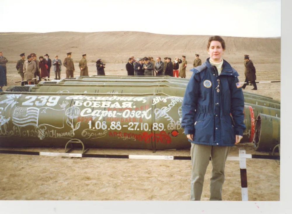 A color photograph ofUS Foreign Service Officer Eileen Malloy in Kazakhstan in 1990 to observe the destruction of missiles banned by the 1988 Intermediate-Range Nuclear Forces Treaty. Malloy is wearing a blue parka and standing alone in front of five Soviet SS-20 intermediate-range ballistic missiles laying horizontally on the ground. Numerous Soviet and US officials stand behind the missiles in the background. The nose cones (which would hold nuclear warheads) have been cut away from the rest of the missiles. The green-painted missile immediately behind Malloy is decorated with graffiti in Cyrillic and drawings of the US and Soviet flags, and includes dates which in context may indicate when the missiles on display were retired under the treaty: August 1, 1988-October 27, 1989.
