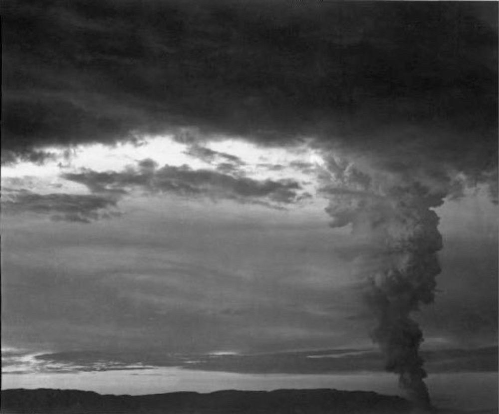 Another black and white photograph of the Trinity explosion taken some minutes after the previous photograph and showing the ominous looking mushroom cloud at a distance, its top spreading out and merging with regular clouds.