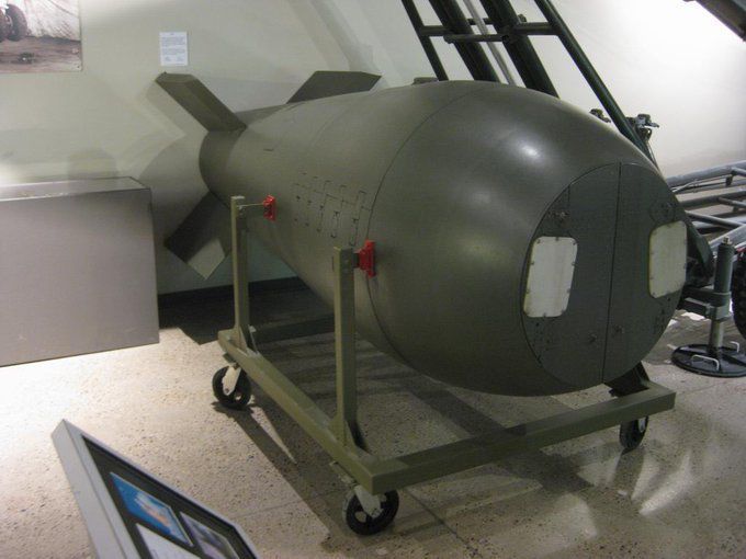 A color photograph of a Mark-5 atomic bomb on display on a wheeled dolly in a museum. The bomb and the cart are painted dark green. The bomb has a blunt nose but otherwise resembles a smaller version of the bulbous “Fat Man” bomb dropped on Nagasaki in 1945.