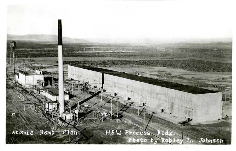 A black and white high-contrast photograph of the T Plant at Hanford, where highly corrosive chemicals were used to separate plutonium from the uranium fuel rods "burned" a nuclear reactor. The building is an 800-foot-long, multi-story windowless rectangle, 65 feet wide and 80 feet tall. There is an even taller stack for venting radioactive emissions in front. Radiation levels inside were so high that nearly all operations had to be carried out remotely. A handwritten caption reads "Atomic Bomb Plant — H.E.W. [Hanford Engineering Works] Process Bldg., Photo by Robley L. Johnson."