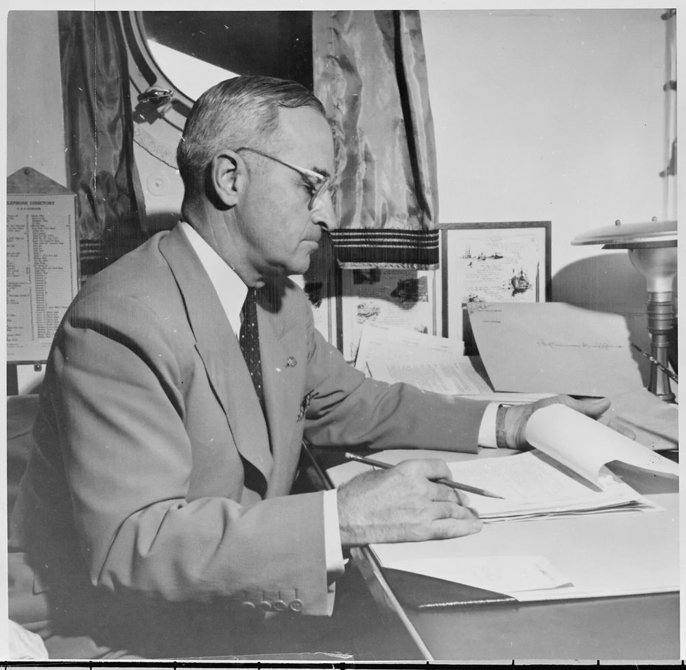 A black and white photograph of President Harry Truman seated at his desk aboard the U.S.S. Augusta while sailing across the Atlantic Ocean to the United States from the Potsdam Conference in Germany, August 6, 1945.