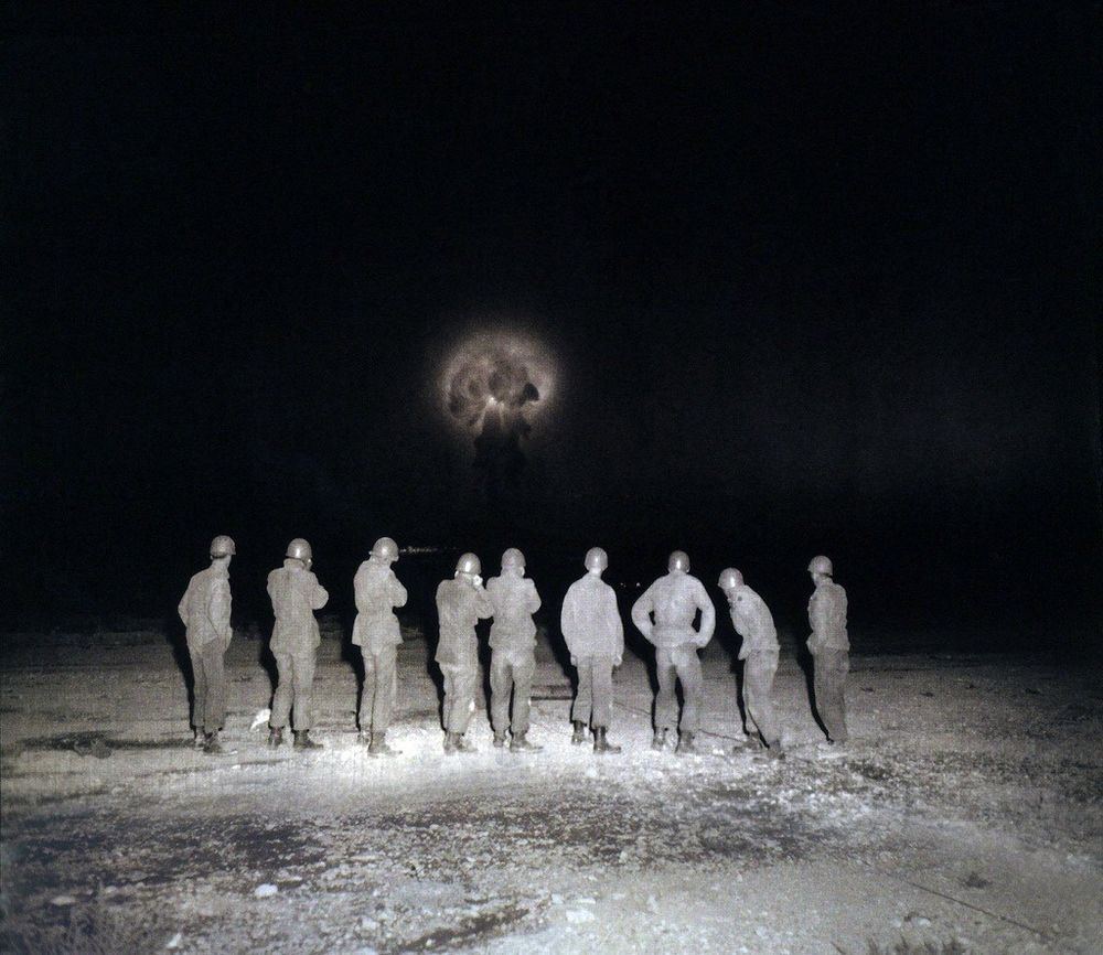 A black and white photograph of the SMOKY test in the same location a few seconds after detonation. Now the nine soldiers are standing and have turned toward the glowing mushroom cloud, which is rising into the sky. They are brightly illuminated by the photographer's flash, while the background—except for the mushroom cloud—is black.