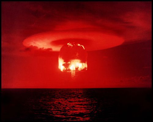 A ground-level color photograph of the large and orange-red glowing ROMEO mushroom cloud rising above Bikini Atoll, with an eerie reddish light reflecting on the ocean in the foreground. The sky is red all around and a circular condensation cloud is forming above the cap of the mushroom cloud.
