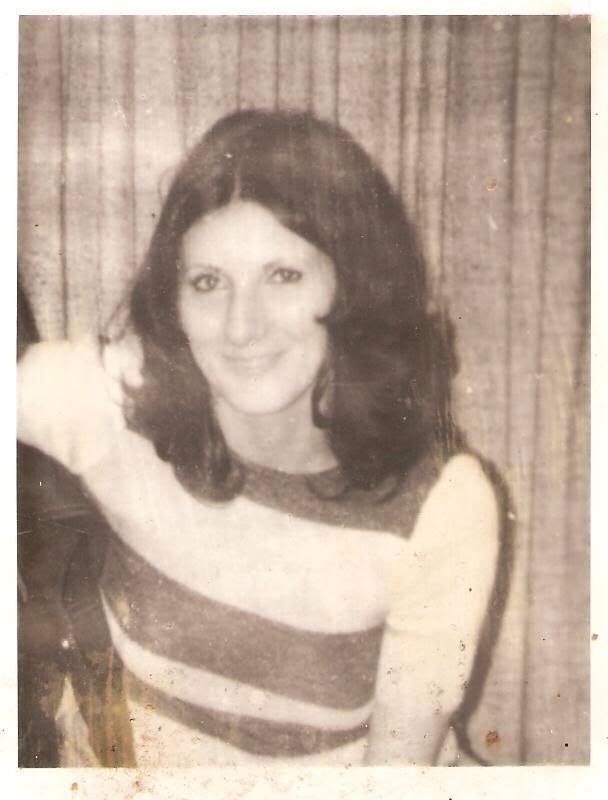 A black and white photograph of Karen Silkwood sitting on a chair in front of a curtain casually posing for the camera. She has long, dark hair and is wearing a striped top.