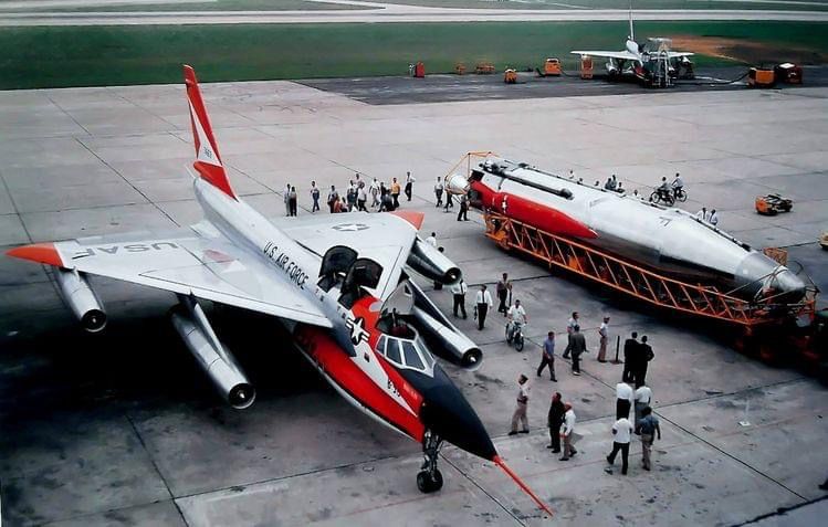 A color photograph showing early versions of the B-58 bomber and the Atlas ICBM (in a wheeled transporter) side by side on a concrete airfield. A few dozen men, most dressed in civilian clothes, a few riding bicycles, are milling around and looking at the weapons. The wingtips, tail, nose spike, and fuel/bomb pod of the bomber are painted bright red.