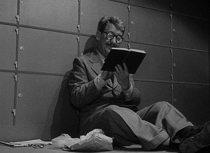 A black and white screenshot from the episode showing Henry Bemis, wearing a light-colored suit and thick eyeglasses, sitting on the floor of the bank vault leaning against the safe deposit boxes and reading a book while eating his lunch.