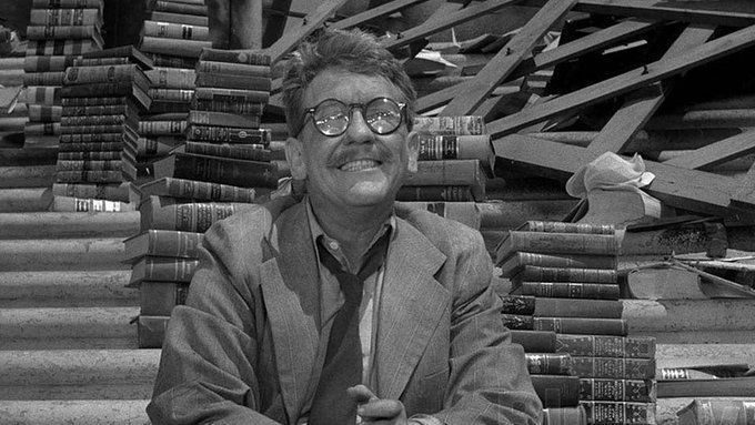 A black and white screenshot from the episode showing Henry Bemis sitting on the steps of the public library, looking up at the sky and smiling, the sun glinting off his thick eyeglasses. He is wearing a light-colored suit, a light-colored dress shirt, and a loosened dark tie. Behind him on the steps are multiple neat stacks of hardcover books.