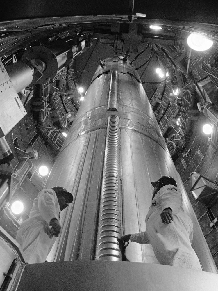 A dramatic black and white photograph looking up from the bottom of a Titan II silo at the missile. Two airman in white coveralls and hard hats are standing at the base of the missile and talking, one of them touching the missile with his right hand.
