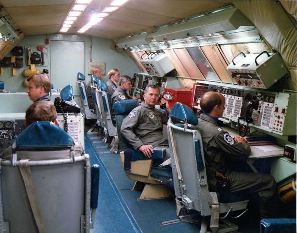 A color photograph of the battle staff area inside "Looking Glass” in the 1980s. The general serving as the Airborne Emergency Action Officer is seated, center, looking at the camera. To his immediate right, an officer wears a holstered revolver strapped to a belt on his flight suit.