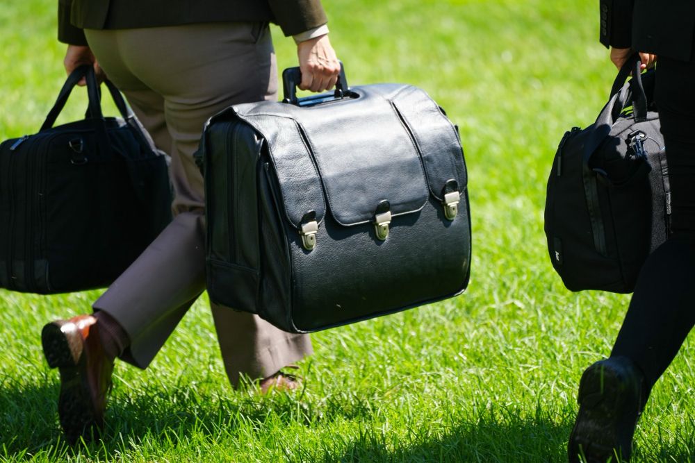 A color photograph showing a close-up look at the distinctive leather-covered Presidential Emergency Satchel being carried across the South Lawn of the White House toward the Marine One helicopter by Lt. Col. Wesmiller.