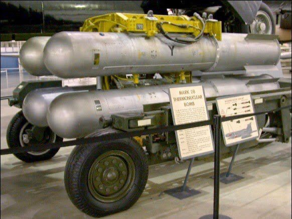 A color photograph of four dull silver, blunt-nosed B28 thermonuclear bombs mounted on a rack attached to a wheeled transport dolly on display in an Air Force museum. Explanatory plaques stand in front of the bombs.