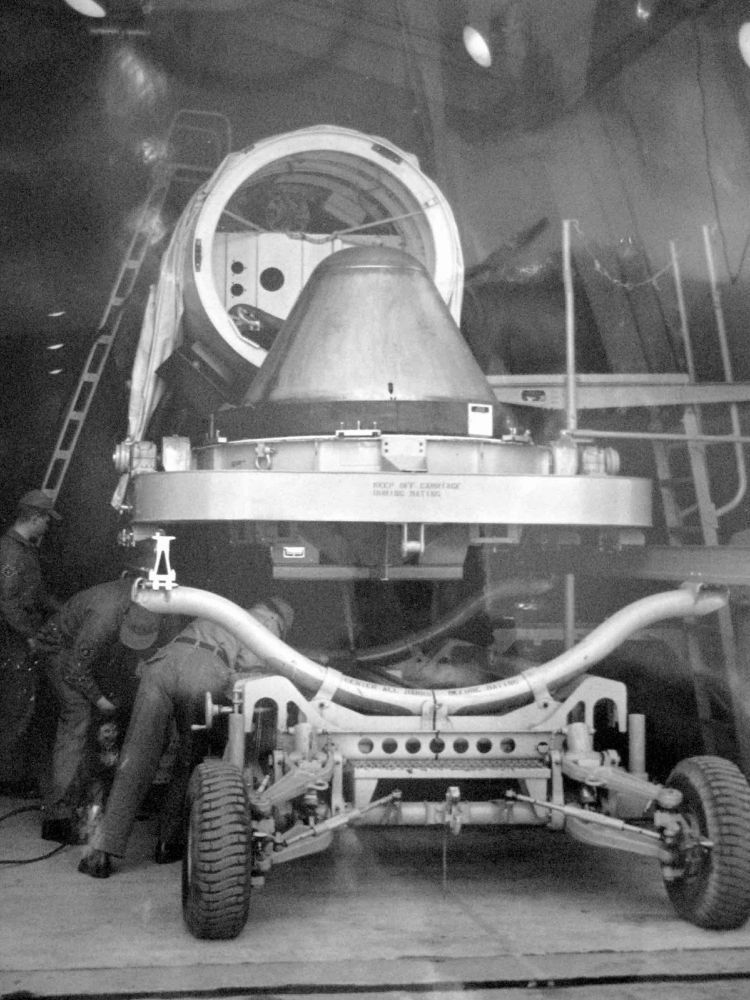 A black and white photograph of a reentry vehicle containing a W49 thermonuclear warhead on a large wheeled transporter prior to being mated to a Thor missile, probably taken at a Royal Air Force base in the United Kingdom in the early 1960s. Three men in fatigues are working below the warhead platform, possibly raising it to the proper height to join the reentry vehicle, which resembles a smooth but slightly squashed thimble, to the missile.