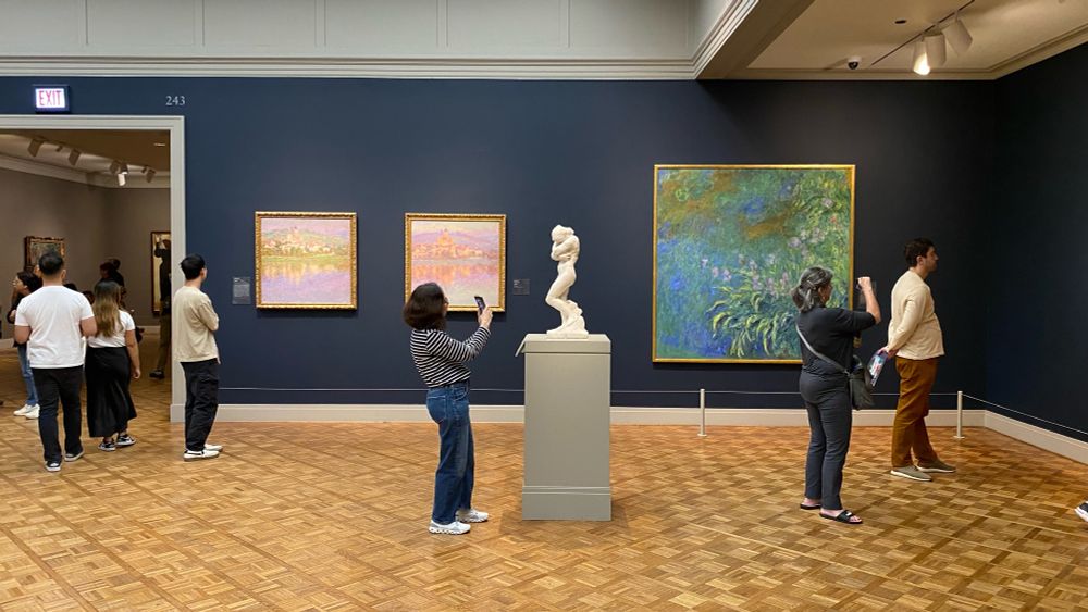 Gallery 243: in by a room filled with many Monets (including his water lilies and wheat stacks series), a woman, center, photographs a small marble sculpture of Eve by Rodin.