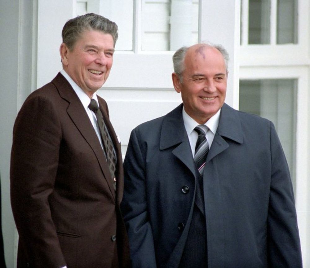 A color photograph of Ronald Reagan and Mikhail Gorbachev posing and smiling for photographers outside Hofdi House in Reykjavik. Reagan is wearing a brown suit and tie, while Gorbachev is wearing a grey overcoat over a grey suit.