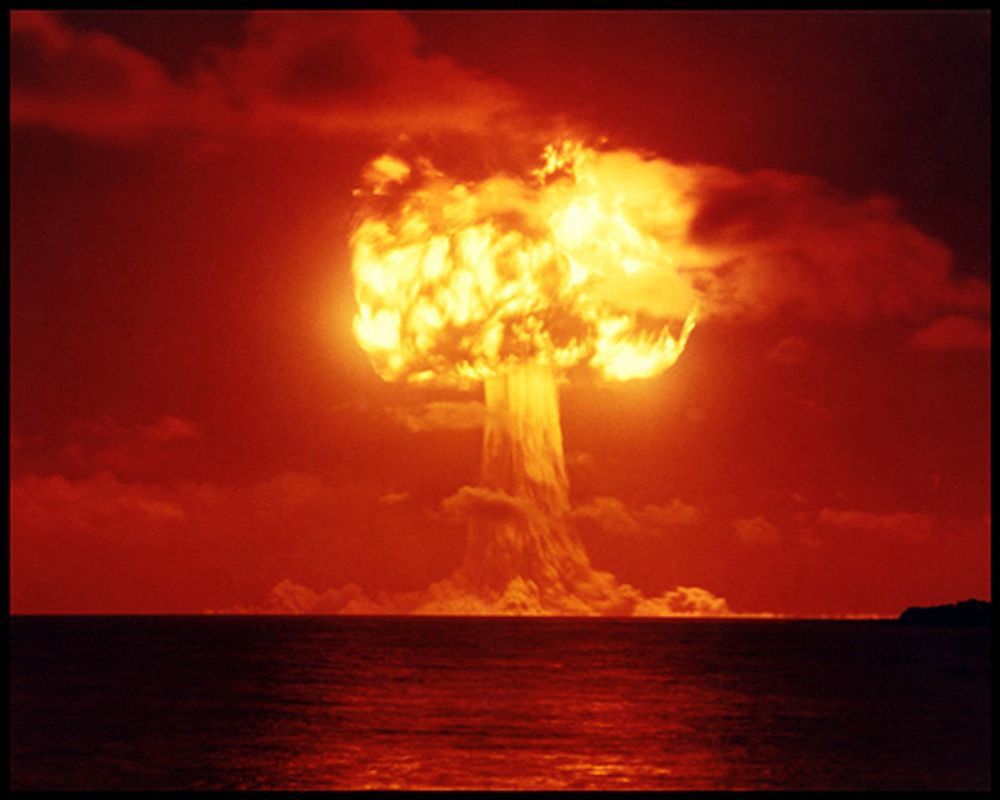 A ground-level color photograph of the very large and glowing mushroom cloud produced by the MOHAWK test of a thermonuclear device on July  2, 1956, on Eberiru Island at Enewetak Atoll. The sky and sea are orange-red and the mushroom cloud is orange-yellow. The MOHAWK device, which weighed 1,116 pounds and was 15 inches in diameter and just over 46 inches long, was detonated atop a 300-foot steel tower and had yield of 360 kilotons. The explosion carved a wide but very shallow crater out of the porous coral soil (1,340 feet wide and 8 feet deep).