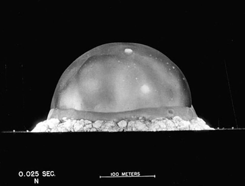 A black and white, high-speed photograph of the Trinity explosion at 0.025 seconds, showing what looks like a very large, slightly irregular bubble with a curtain of dust all around its bottom edge against a pitch dark sky. A scale indicating “100 meters” shows that the fireball was already approximately three times that size.