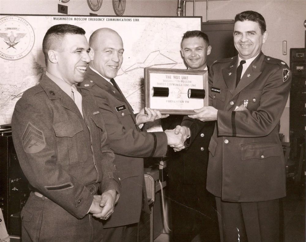 A black and white photograph circa August 1963 commemorating the establishment of the Direct Communications Link. Four white male officers are smiling and standing in what is probably a room in the National Military Command Center, two of whom are shaking hands and holding a presentation plaque on which two pieces of the undersea cable used to transmit messages between Moscow and Washington are affixed. A map in the background shows the path of the cable between the two national capitals.