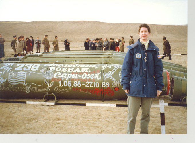 A color photograph of US Foreign Service Officer Eileen Malloy in Kazakhstan in 1990 to observe the destruction of missiles banned by the 1988 Intermediate-Range Nuclear Forces Treaty. Malloy is wearing a blue parka and standing alone in front of five Soviet SS-20 intermediate-range ballistic missiles lying on the ground. Numerous Soviet and US officials stand behind the missiles in the background. The nose cones (which would hold nuclear warheads) have been cut away from the rest of the missiles. The green-painted missile immediately behind Malloy is decorated with graffiti in Cyrillic and drawings of the US and Soviet flags, and includes dates which in context may indicate when the missiles on display were retired under the treaty: August 1, 1988-October 27, 1989.