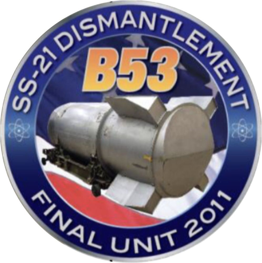 An image of a circular logo patch designed by the National Nuclear Security Administration to commemorate the dismantlement of the last B53 thermonuclear bomb in October 2011. "SS-21 Dismantlement" is written in silver capital letters on the top edge of the circle (referring to an extensive "start-up" activity that precedes any actual dismantlement work at the Pantex Plant in Amarillo, Texas). Stylized atomic symbols (also silver) appear before and after these words. "Final Unit 2011" is written in silver capital letters along the bottom edge. In the center is "B53" in large yellow letters/numbers above a circular photo of a B53 on a dolly similar if not identical to the one in the first post, with a background showing a portion of the American flag (white stars on a blue field, red and white stripes).