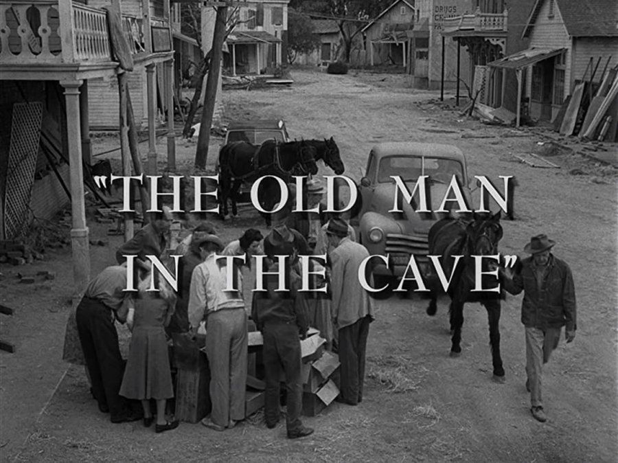 A black and white screenshot from the episode showing the title over a shot of an unpaved street in ramshackle Western-looking town. In the foreground, a small group of men (all wearing hats) and women (all wearing dresses) are gathered around a pile of cardboard boxes. Next to them, a man leads a black horse pulling a car.