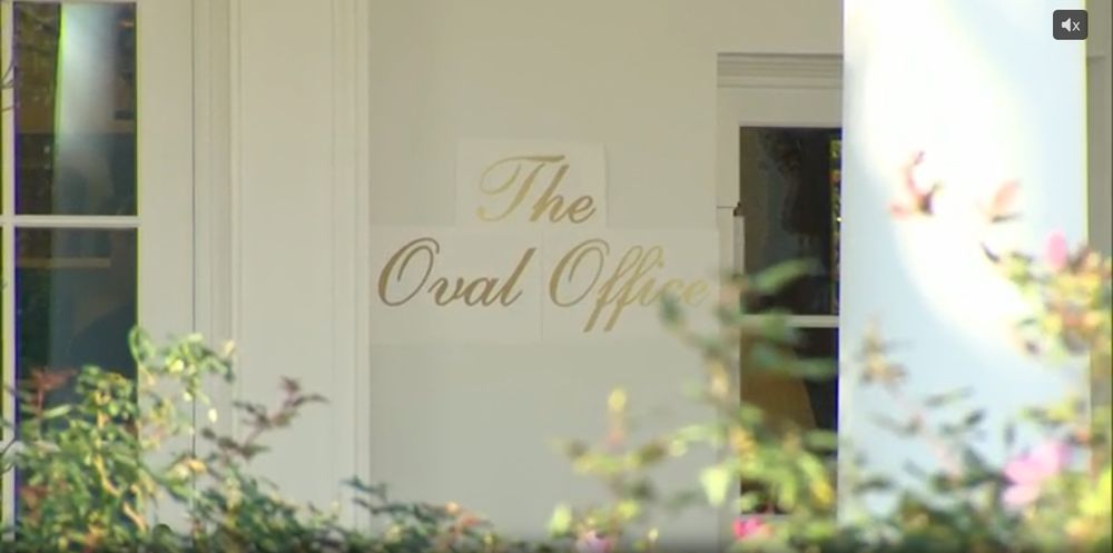 A screenshot of the door to the Oval Office, next to which are the words, in large gold script, "The Oval Office," on three pieces of paper attached to the exterior wall. Not visible, two large gold appliqués Trump has installed above the door (similar to what he's attached to the walls of the Oval Office and the Cabinet Room.