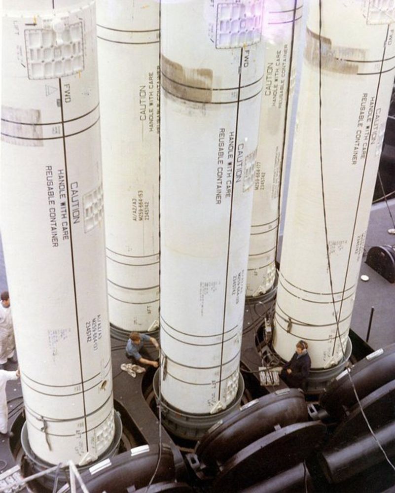 A color photograph of five Polaris A3 submarine-launched ballistic missiles in reusable white cylindrical containers being loaded into the launch tubes of a Lafayette-class submarine. Two sailors can be seen next to the launch tubes of two of the missiles, providing a good indicator of their significant size.