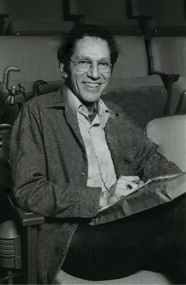 A black and white portrait of Tom Lehrer sitting in a seat in a chair in a theatre circa 1983.