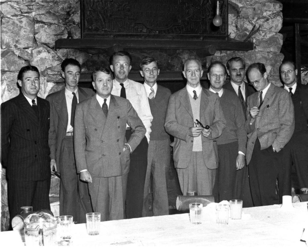A black and white photograph of the members of the Office of Scientific Research and Development posing in front of a large stone fireplace during a meeting at the Bohemian Grove in Monte Rio, California, in September 1942. J. Robert Oppenheimer, who was not a member but was advising the OSRD’s S-1 Executive Committee, is second from the left.
