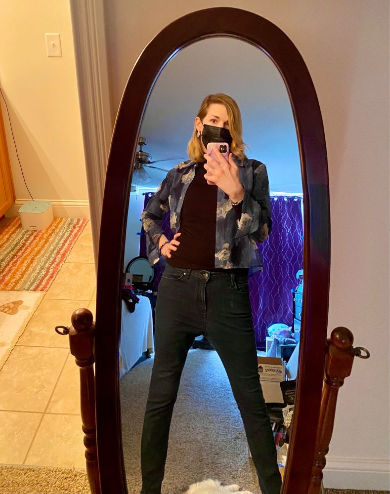 Masked blonde trans woman failing to boy mode by wearing skinny jeans, a black skin tight title neck and a translucent floral blouses as an accent. Striking a wide stance in an oval mirror